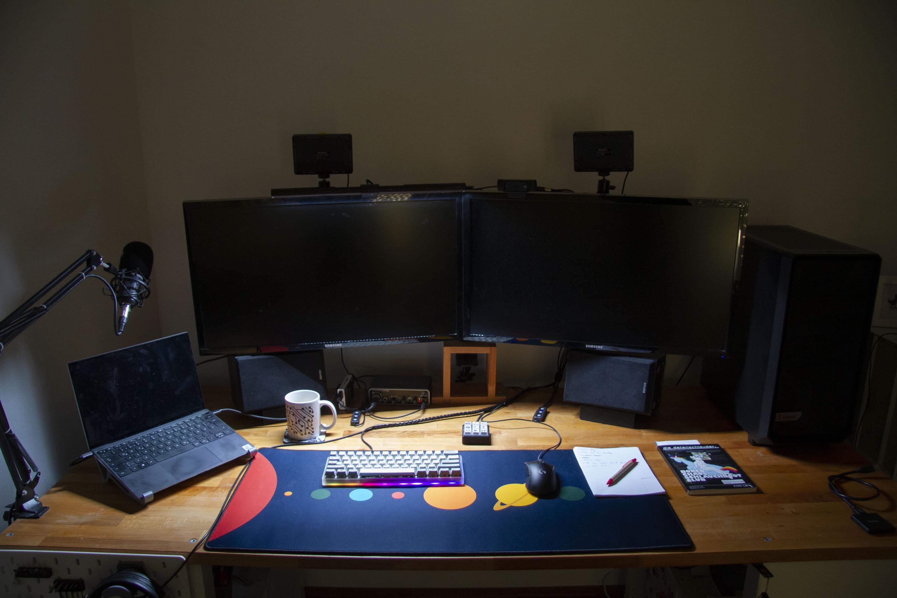 Foto von vorne von meinem Schreibtisch im abgedunkelten Zimmer. Links steht ein Laptop, dann zwei große Monitore, dann ein Desktop gehäuse. Die Screenbar hängt auf dem linken Monitor und beleuchtet den Schreibtisch. Auf dem Schreibtisch liegt eine blaue Deskmat mit bunten Planeten und darauf ein kleines graues Keyboard, eine schwarze Maus und ein Notizzettel mit Kugelschreiber. Unter den Monitoren stehen 2 Boxen, eine Kaffeetasse und ein Audiointerface.